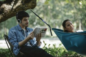 Delighted young multiethnic couple in casual clothes reading book and resting in hammock while spending time together in nature